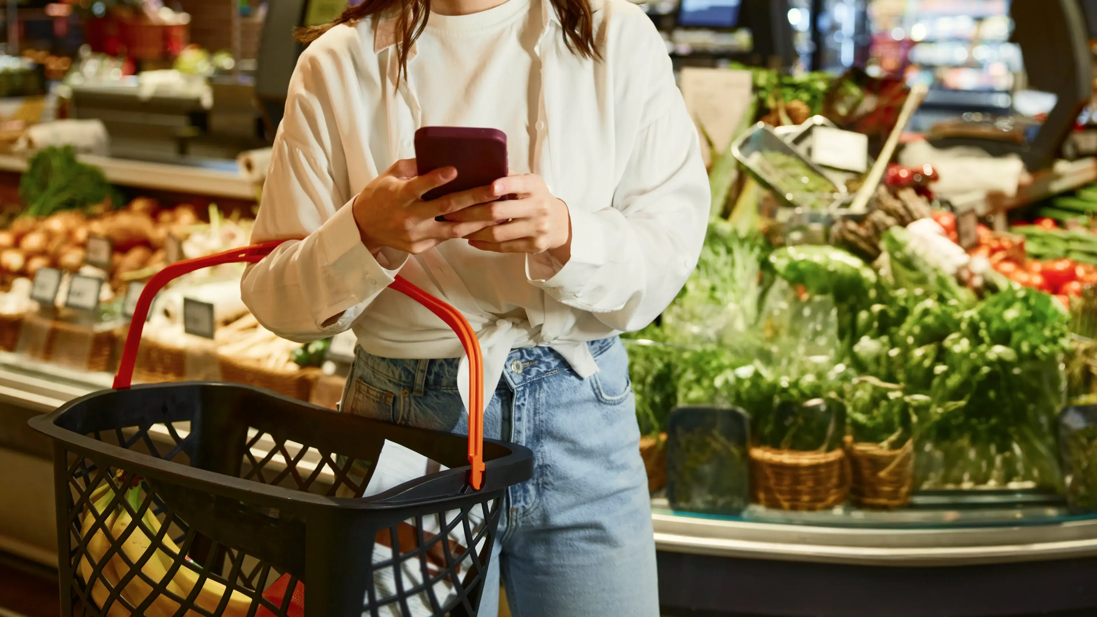 Lekkerland Shopper checking smartphone in supermarket produce aisle, holding black basket with red handles. Retail lifestyle photography