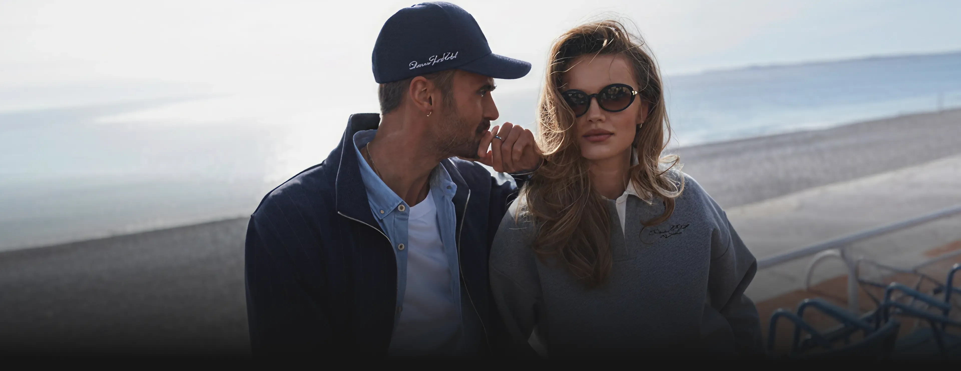 Diverse Sport Club SS26 lookbook couple on seaside promenade navy cap and jacket grey logo sweatshirt candid style